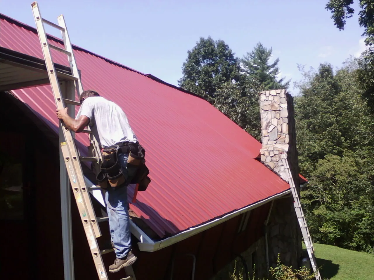 Roofer on a ladder beside a red metal roof and stone chimney — Soffit & Fascia Repair by Roofing Brothers in Forman